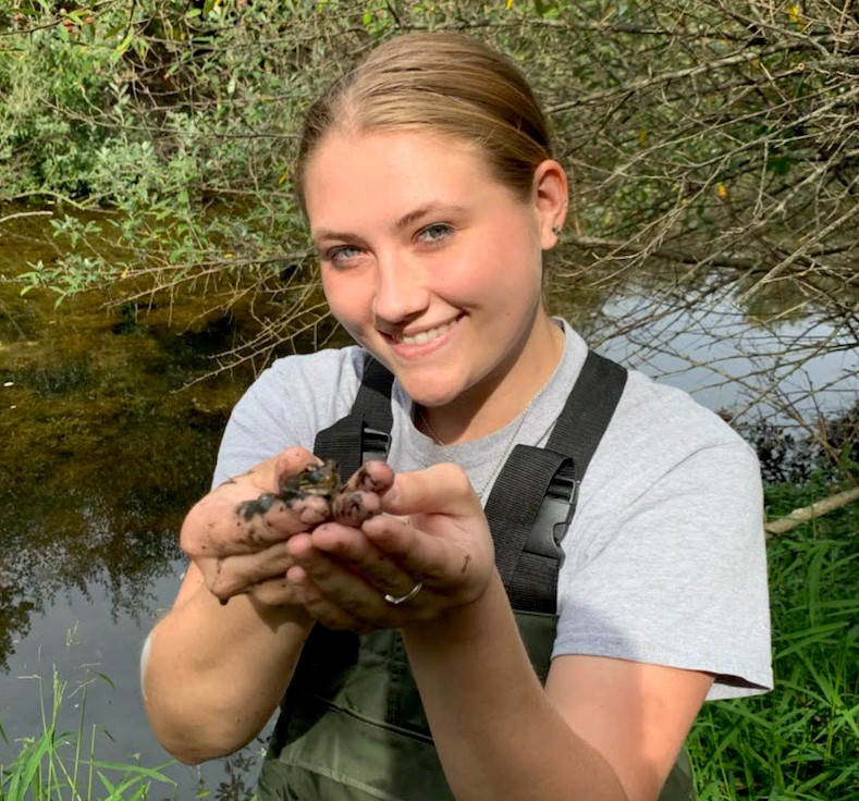Brianna Sander smiles at the camera and holds up a tiny frog