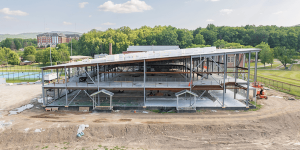 Aerial view of a building under construction with a steel framework, adjacent to an existing brick building, surrounded by construction materials and trees.