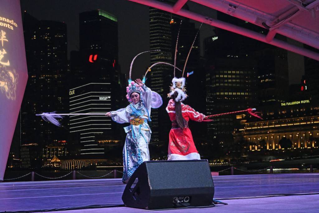 Two performers in elaborate traditional Chinese opera costumes engage in a stylized combat scene on an outdoor stage at night. One wears blue and white armor, the other red, both holding long spears with flowing tassels. The city skyline with illuminated skyscrapers and the Fullerton Hotel glows in the background.
