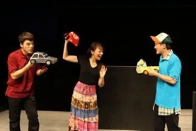 Three actors perform a lively scene on stage. Two hold toy cars and appear to be playfully arguing, while the third, standing between them, reacts with exaggerated surprise. The simple black background emphasizes their colorful costumes and expressive gestures, suggesting a comedic or educational theatre piece.