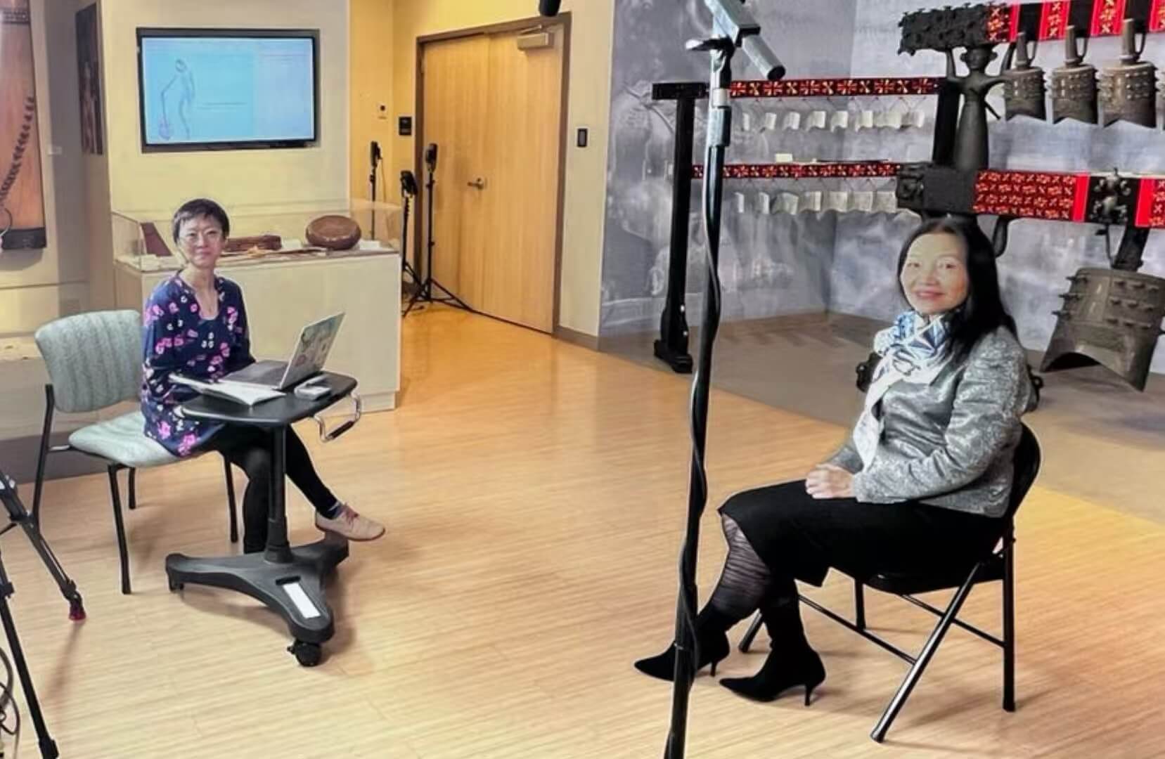 Two professors sit facing each other in the exhibit area of the center during the interview session. Professor Song operates a laptop, while Professor Mei Han sits across from her under studio lights. Behind them are traditional Chinese musical instruments, including bronze bells and zithers, displayed against an elegant backdrop.