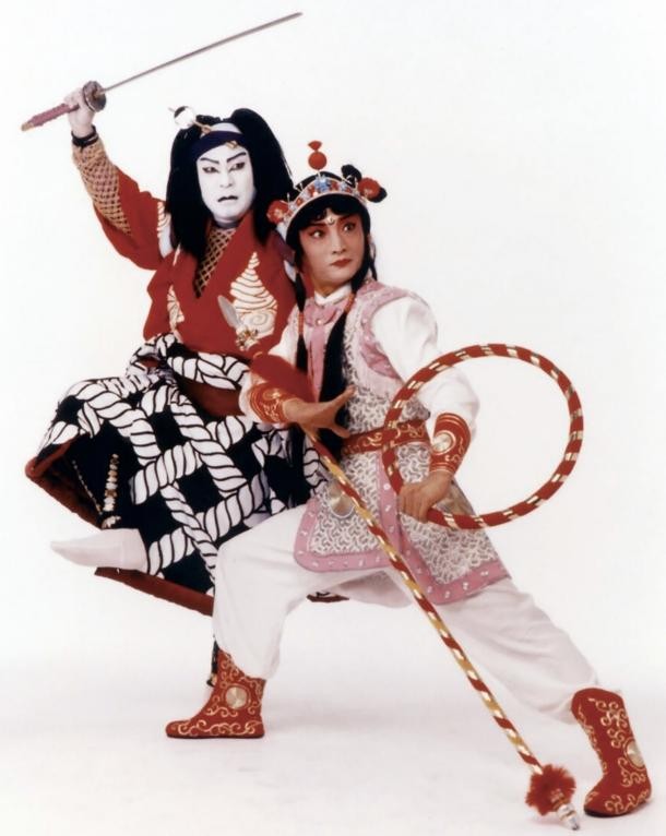 Two performers in traditional theatrical costumes pose dramatically against a plain background. Ichikawa Ennosuke III wears red and black attire with bold white face paint and wields a sword, while the other, Li Guang, dressed in pink and white armor-like costume, holds a staff in a martial arts stance.