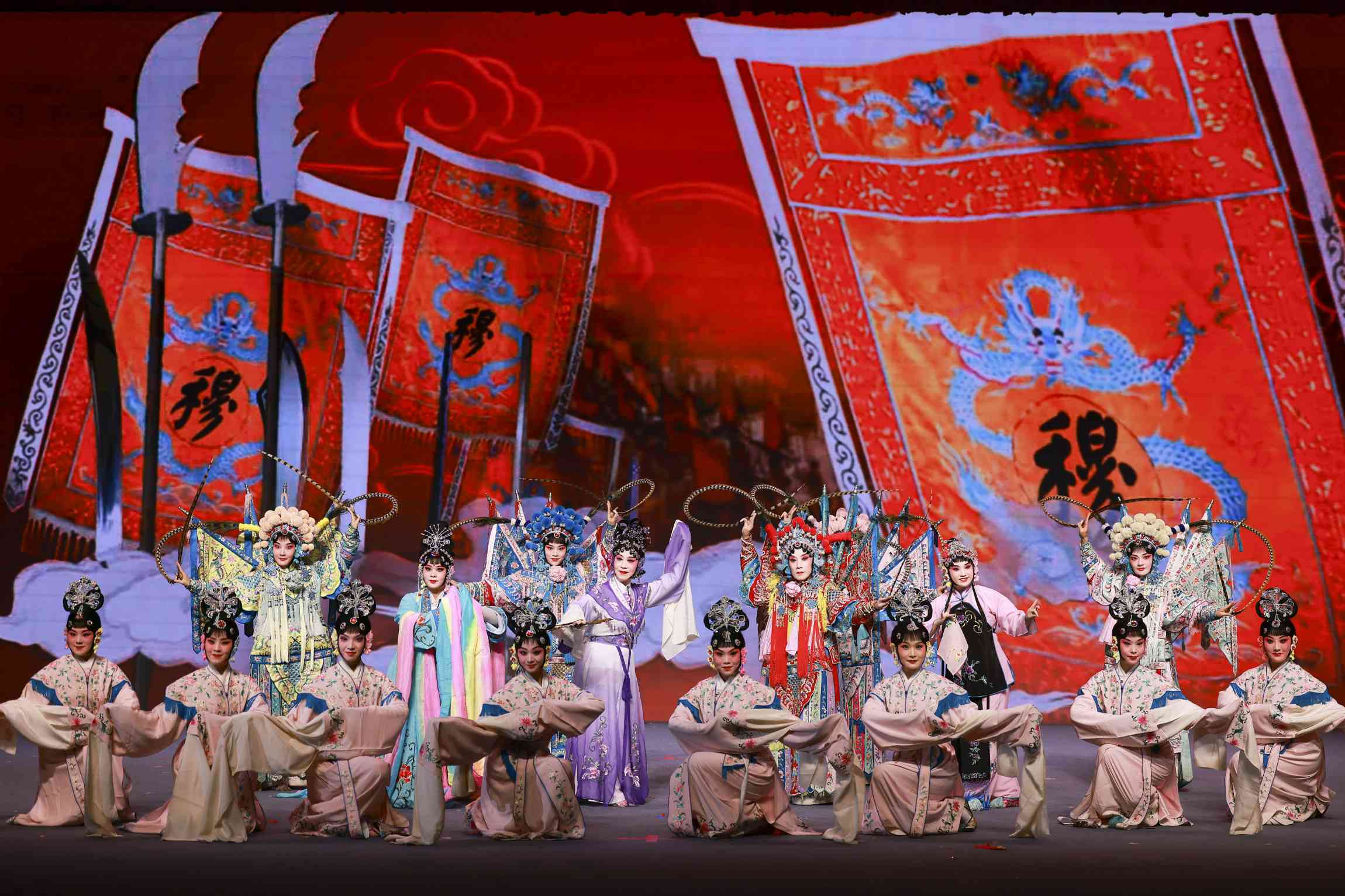 A large group of performers in elaborate traditional Chinese opera costumes pose on stage. The front row features dancers in elegant pastel robes, while the back row showcases warriors in ornate armor with feathered headdresses. Behind them, a vivid red backdrop displays large banners with dragons and Chinese calligraphy, creating a grand and dramatic atmosphere.