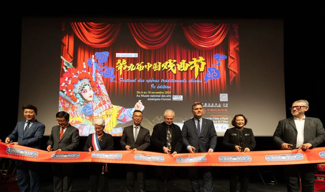 A group of officials and guests stand on stage cutting a ceremonial ribbon at the opening ceremony. Behind them, a large screen displays colorful Chinese opera artwork and event details in Chinese and French, set against a red curtain background.