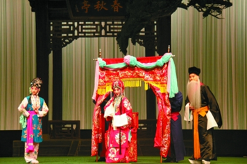 Performers in traditional Chinese opera costumes act out a scene on stage. One performer sits inside a decorated red sedan chair while others stand around in colorful robes and headpieces. The backdrop features a pavilion structure and hanging curtains, creating an elegant classical setting.