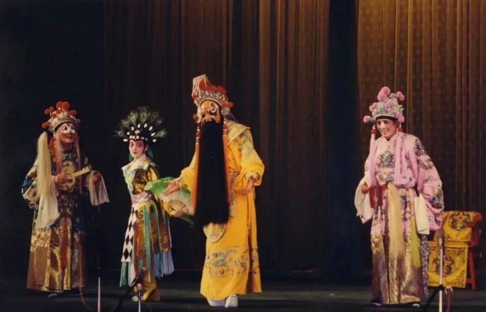 Four performers in elaborate traditional Chinese opera costumes stand on stage. The central figure, dressed in a golden robe with a long black beard, gestures toward others in colorful embroidered attire and ornate headpieces. The scene unfolds against a simple curtain backdrop, highlighting the characters’ expressive poses and costumes.