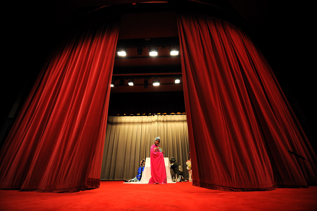 A Kunqu performer in traditional Chinese opera costume stands center stage under dramatic lighting, framed by large red velvet curtains. The stage floor and curtains create a rich, theatrical atmosphere, while additional performers in colorful costumes are visible in the background near the gray backdrop.
