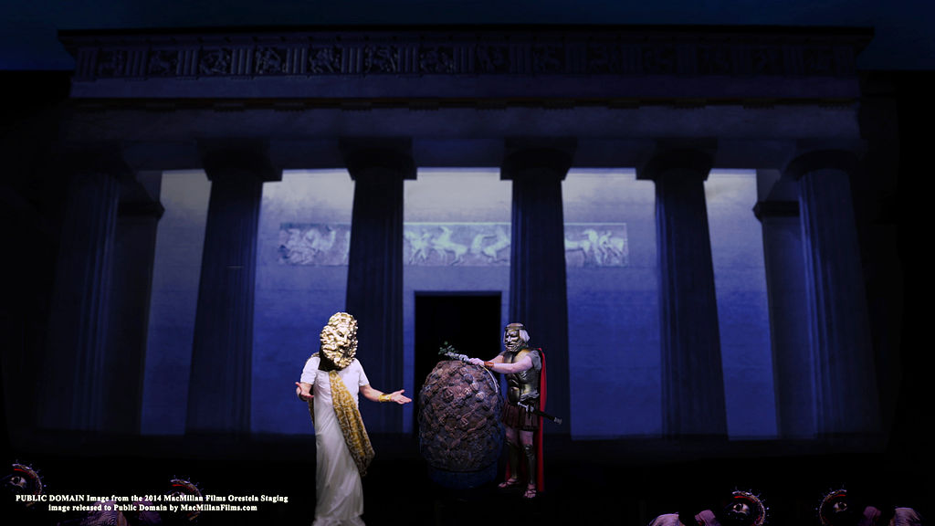 Two actors perform in front of a large projection of an ancient Greek temple façade. One wears a white robe and golden mask resembling a mythological figure, while the other, dressed as a soldier in armor and a red cape, holds a large round shield. The lighting creates a dramatic, classical atmosphere.