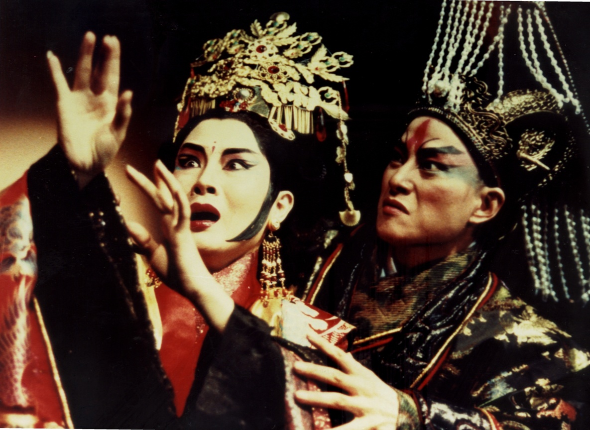 Two performers in traditional Chinese opera costumes act out an intense moment on stage. The figure on the left, wearing a gold headdress and elaborate makeup, looks startled with raised hands, while the other, dressed in dark brocade with a fierce expression, grips her arm.