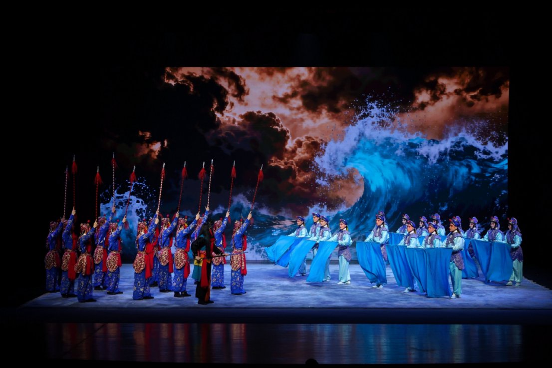 京剧《白蛇传·水斗》舞台演出场景,演员分列舞台两侧进行对峙。左侧演员身着蓝色铠甲戏服,高举红缨长枪;右侧演员穿着浅蓝色戏服,挥动长幅蓝绸模拟翻涌水浪。背景大屏幕呈现巨浪与乌云翻腾的海天景象,营造出激烈壮观的水战氛围。