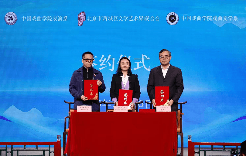 三位代表站在铺有红布的桌子后,在一场战略合作签约仪式上高举红色签约本。蓝色的背景墙上印有中国戏曲学院表演系、戏剧文学系以及北京市西城区文学艺术界联合会的标志。