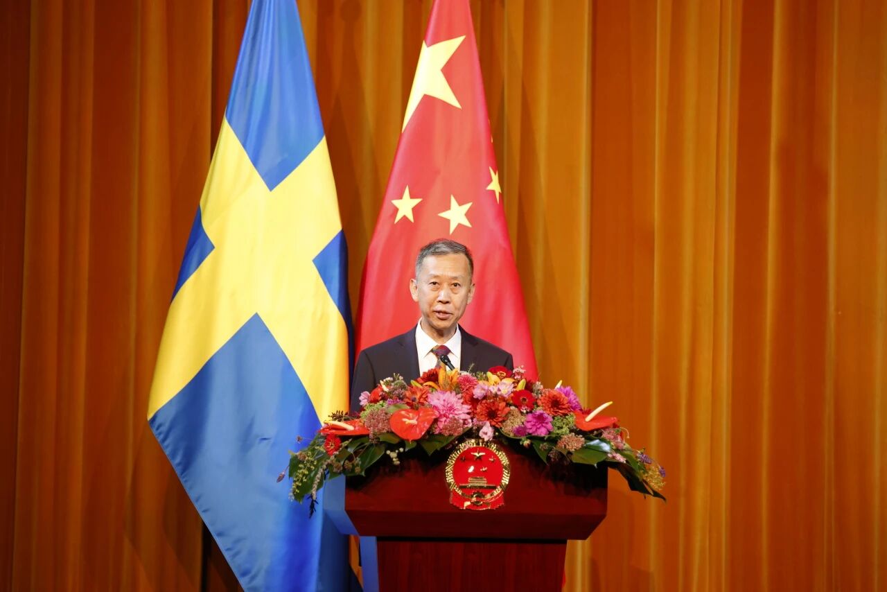 Cui Aimin stands at a podium decorated with colorful flowers, delivering a speech in front of Swedish and Chinese national flags.
