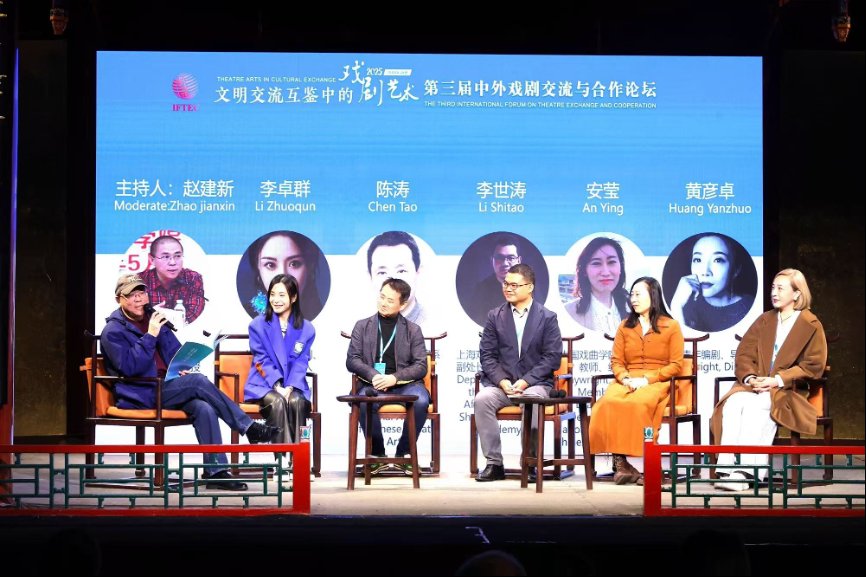小组座谈会舞台全景照片,六位演讲嘉宾并排坐在中式传统木椅上。身后的蓝色电子大屏幕上印有活动名称“第三届中外戏剧交流与合作论坛”,以及各位嘉宾的头像和姓名:主持人赵建新、李卓群、陈涛、李世涛、安莹和黄彦卓。最左侧身穿深色夹克、戴着棒球帽的主持人正拿着手册并对着麦克风发言。