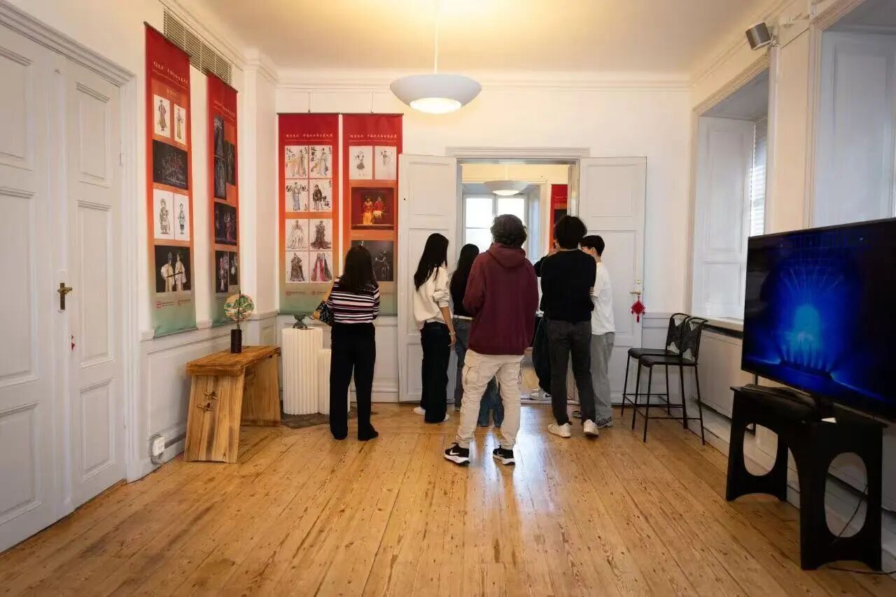 Visitors walk through a gallery exhibition featuring vertical banners displaying Chinese opera costumes, character designs, and stage photography. The bright room has wooden floors, white walls, and a television screen showing blue stage lighting imagery on the right side. Several attendees stand and observe the exhibits in the adjoining room.