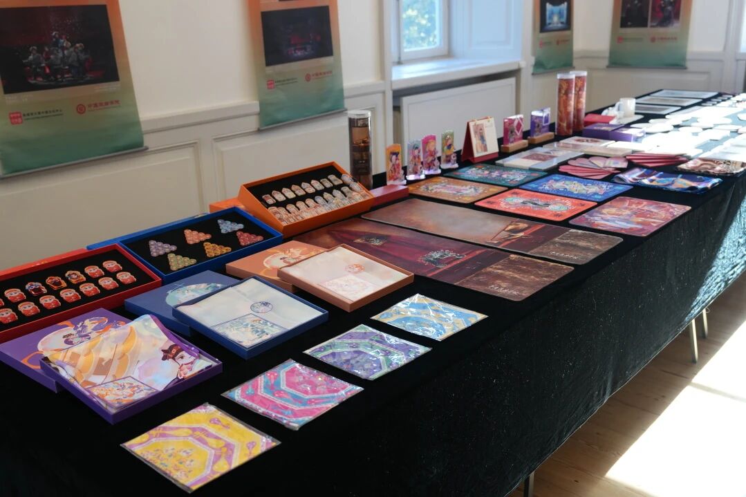 Display table featuring colorful Chinese opera–themed merchandise and exhibition materials arranged on a black tablecloth in a gallery space. Items include illustrated cards, pins, notebooks, posters, bookmarks, and decorative boxes inspired by Chinese xiqu stage art.
