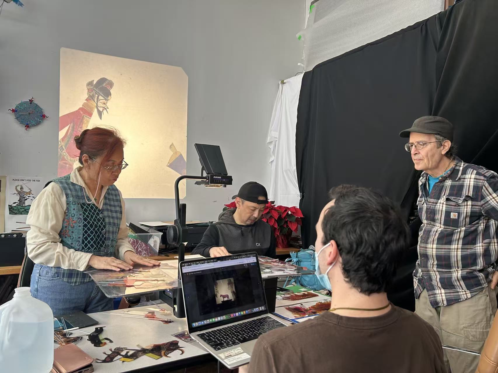 A group of four people working in an art or media studio, examining traditional Chinese shadow puppets. A woman on the left carefully handles a puppet under a document camera, while a laptop in the foreground displays a digital preview of the artwork.