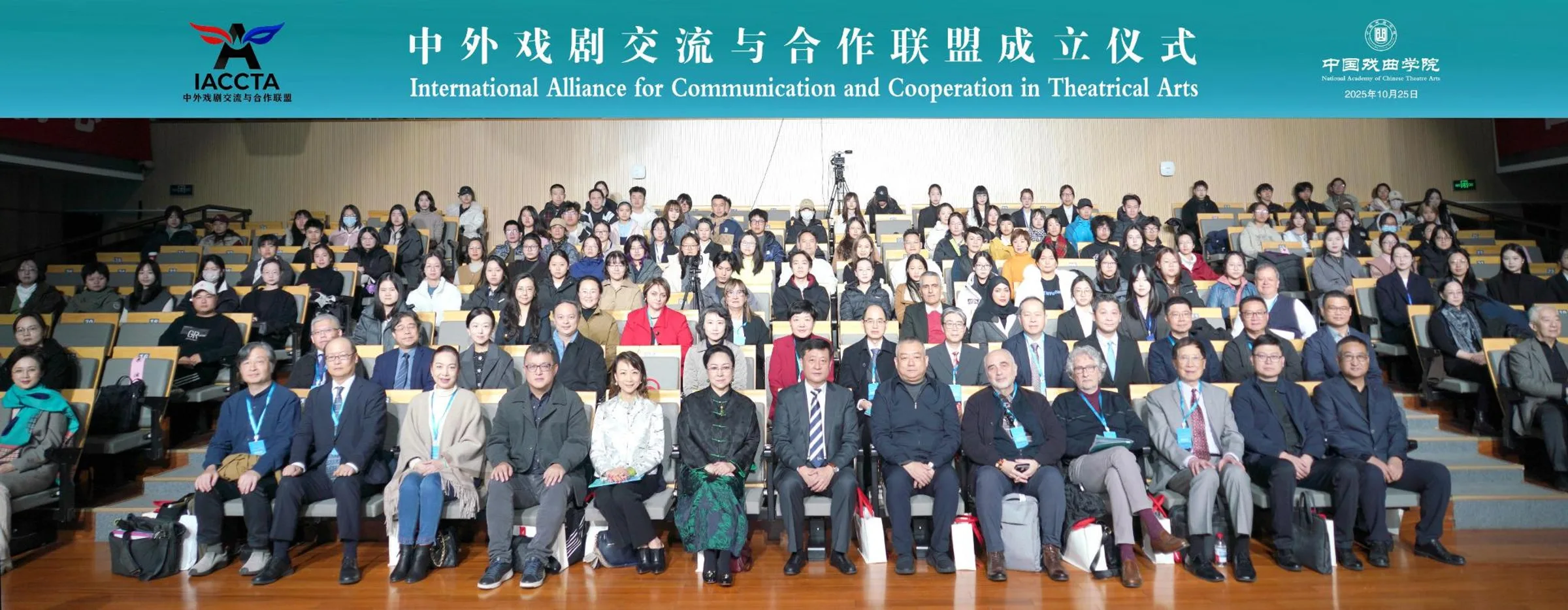 中外戏剧交流与合作联盟成立仪式的大合影。众多参会人员在礼堂的阶梯座位上合影留念。画面顶部有一条青绿色的横幅,上面印有中英文活动名称,日期为2025年10月25日。