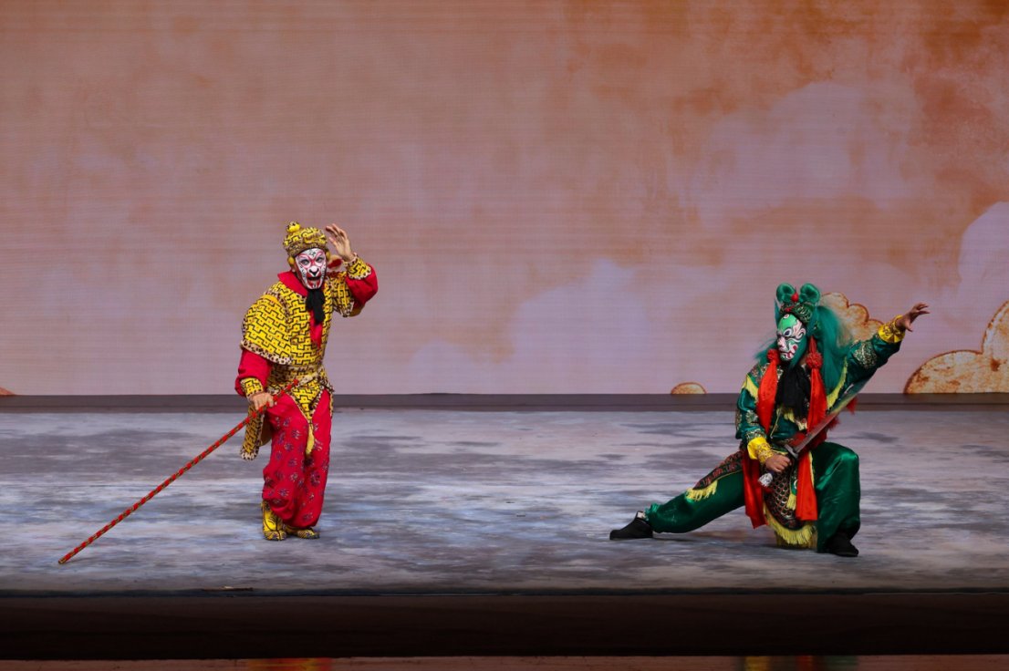 两位身着《大闹天宫》主题戏服的中国戏曲演员在舞台上表演。左侧演员穿着红黄相间、带有孙悟空脸谱妆容的戏服,手持长棍并做出张望动作;右侧演员身着绿色戏服,半蹲持兵器摆出武打姿势。