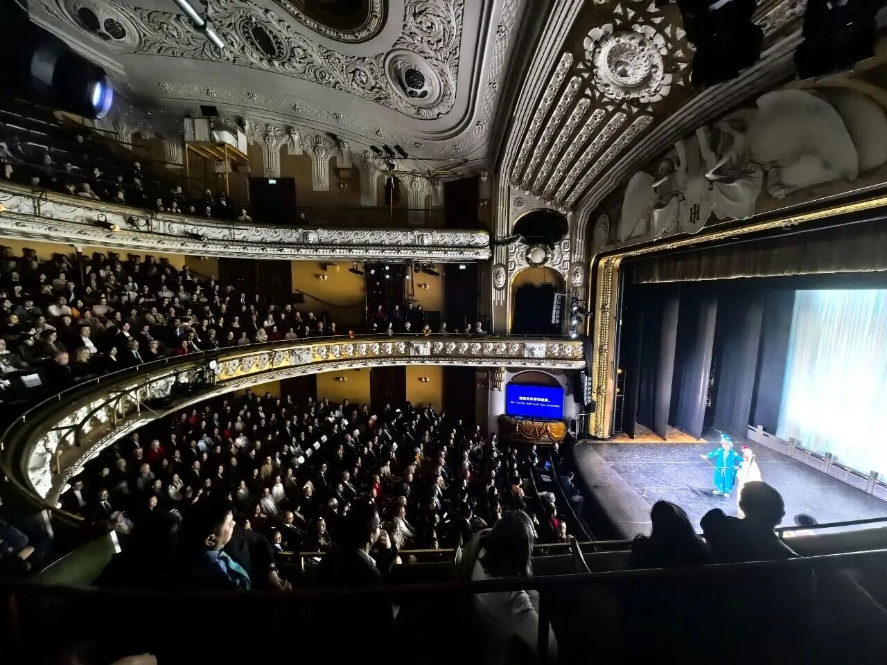 Wide view of the Oscar Theatre filled with audience members seated across multiple balcony levels during a live stage performance. Two performers in brightly colored costumes stand on the stage. 