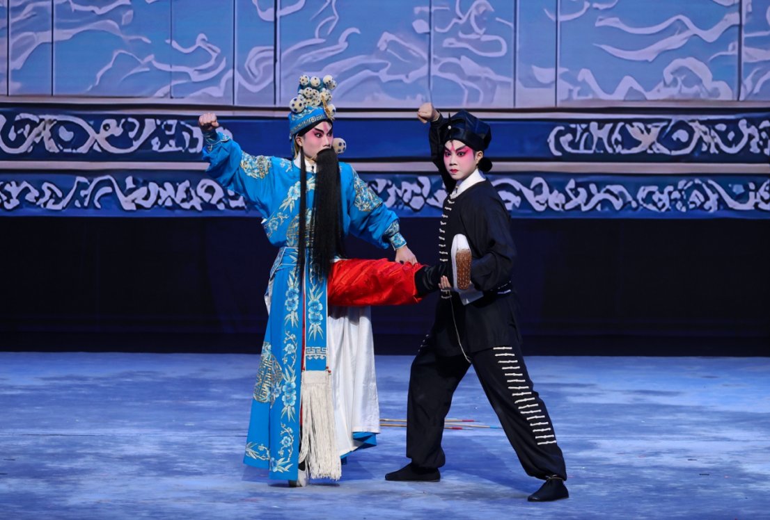 两位演员在舞台上表演粤剧片段《马福龙之卖箭》。左侧演员身穿蓝色绣花官服,头戴华丽头饰并留长黑须;右侧演员身着黑色戏服