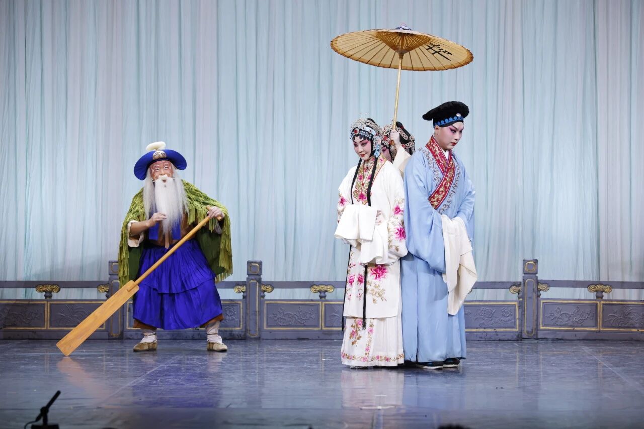 A stage scene from a Chinese opera featuring three performers. On the left, an older male character wearing a blue hat, green cape, and blue robe poses dynamically with a wooden boat paddle. On the right, a female character in an elegant white robe with pink floral embroidery stands back-to-back with a male character in a light blue robe, sheltered under a traditional paper umbrella.