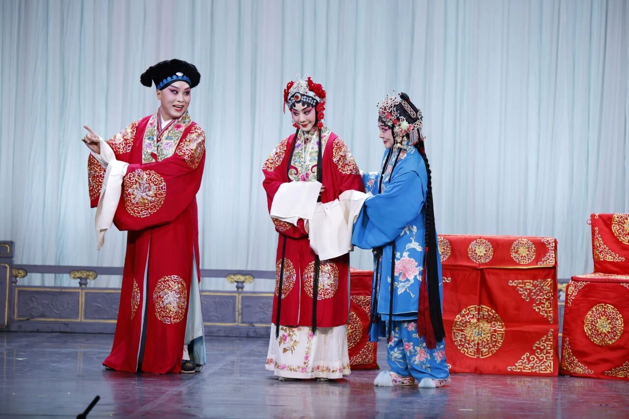 Three Chinese opera performers on a stage decorated with red ceremonial furniture. On the left, a male character in a red robe points forward. In the center, a female character in a matching red patterned robe holds hands with another female character on the right, who is dressed in a vibrant blue embroidered robe.