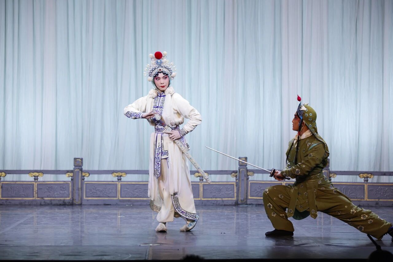 A martial arts scene from a Chinese opera. A female warrior character in a white and blue outfit with a feathered headpiece poses gracefully with a sheathed sword. On the right, an opponent in olive-green military attire crouches in a low stance, pointing a straight sword toward her.