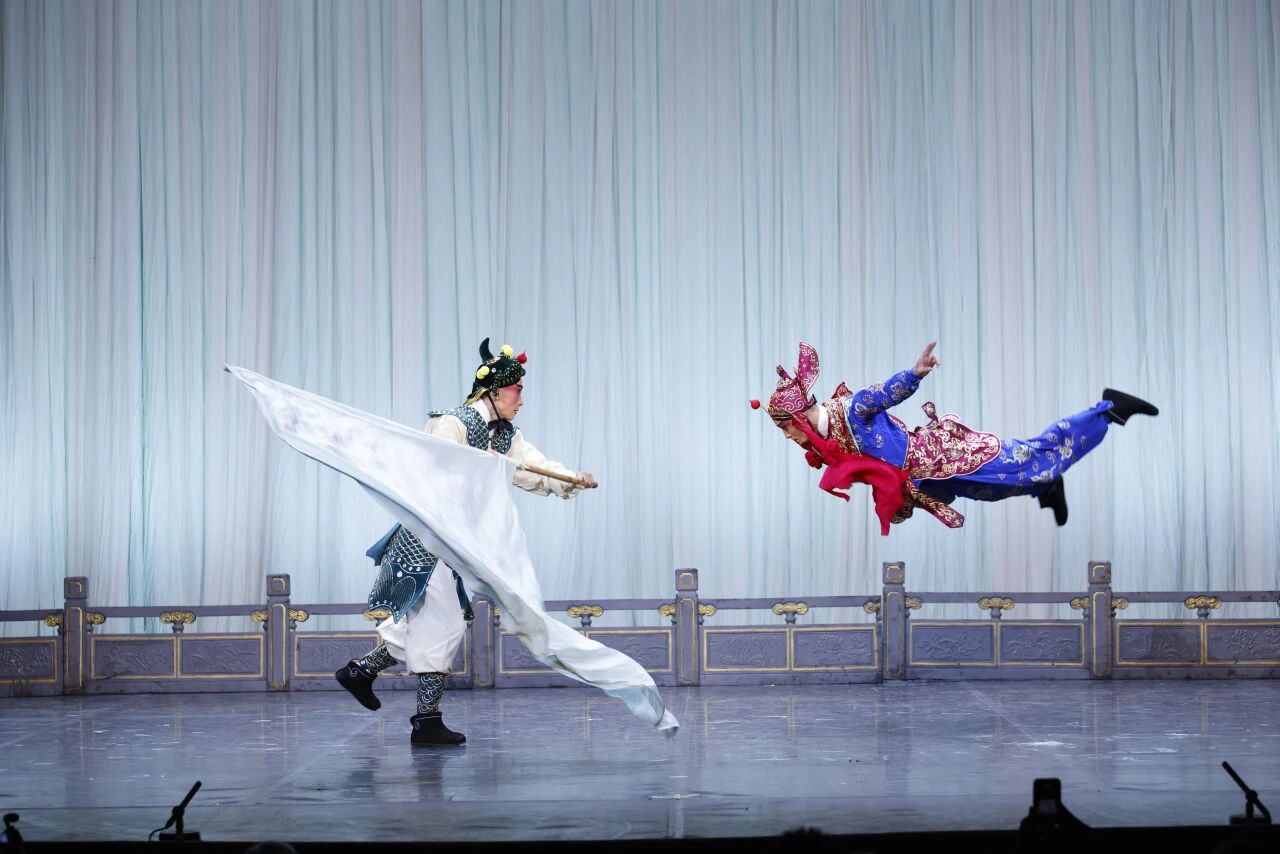 An action-packed Chinese opera scene featuring two acrobatic performers. On the left, a character in green armor sweeps a large white flag horizontally. On the right, an acrobatic performer in a blue and red outfit is captured mid-air, flying horizontally across the stage.
