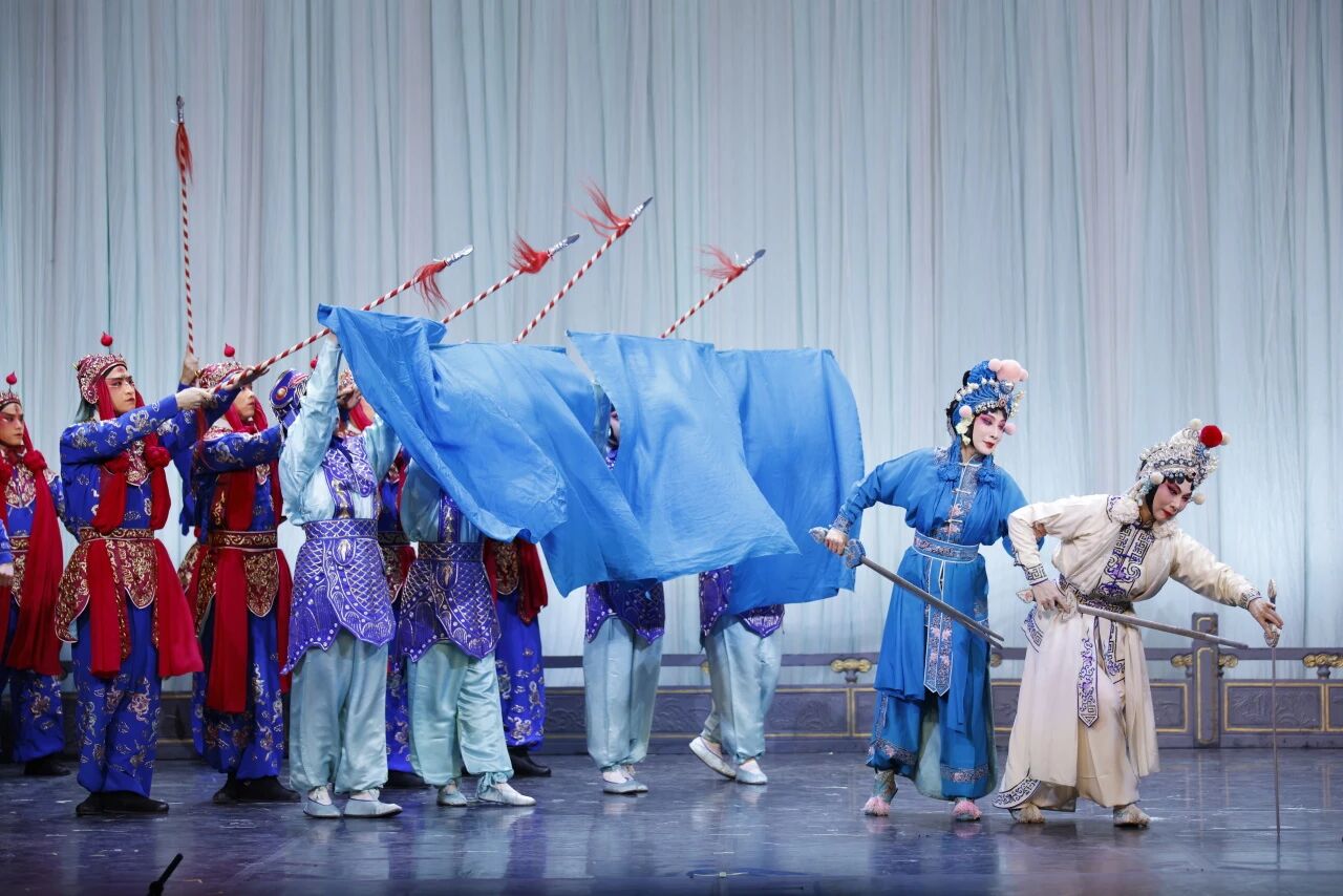 A large ensemble martial scene from a Chinese opera. On the left, multiple soldiers in blue and red uniforms hold up spears and large blue banners representing water or waves. On the right, two female warriors—one in blue and one in white—are locked in a dynamic sword fight in the foreground.