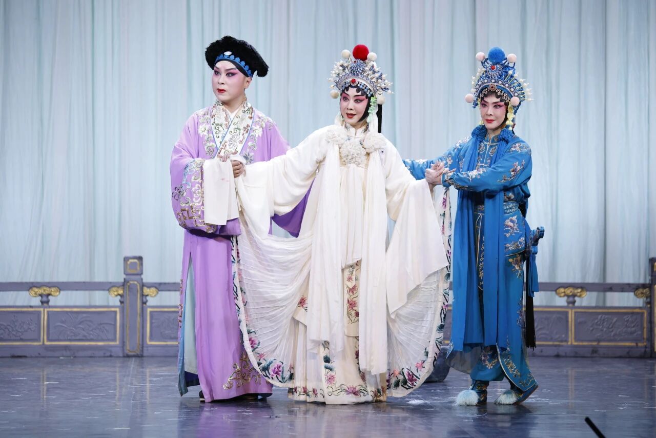 A dramatic final pose of three Chinese opera characters. In the center, a female character in an elaborate white dress with floral details extends both arms. She is flanked and supported by a male character in a purple robe on her left, and another female character in a blue robe on her right.