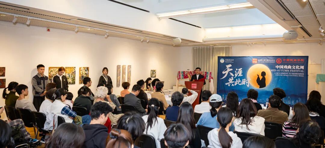 Audience members seated in a gallery-style space listen to a speaker at a podium during Chinese Opera Cultural Week. A large blue bilingual banner behind the speaker features Chinese and English text about the festival and a silhouette of a person against a moonlit background. Paintings are displayed on the walls, and several staff members stand along the side of the room.
