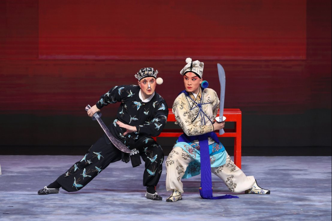 Two performers pose onstage during a Chinese opera performance, each holding a curved sword in a martial arts stance. One performer wears a black costume with blue embroidered patterns, while the other wears a white and light blue embroidered costume with traditional opera makeup.