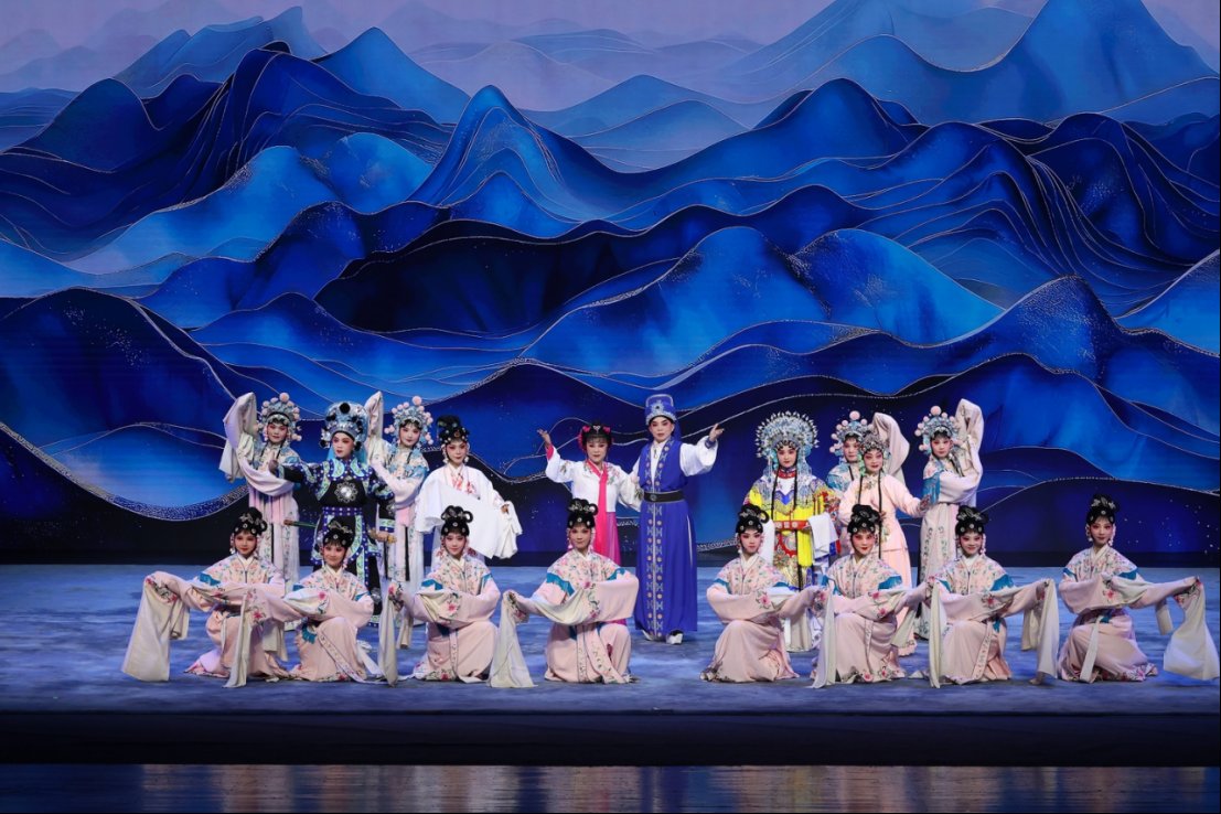 多位身着华丽传统戏曲服饰的演员在舞台上集体亮相,表演《潮闽琼韵·狮城交响》。演员分前后两排排列,前排演员身穿浅粉色戏服,手持长水袖;后排演员则穿着不同剧种特色的蓝色、白色和彩色戏服与头饰。舞台背景以层叠起伏的深蓝色山峦图案构成,营造出壮丽而富有东方美学的视觉效果。