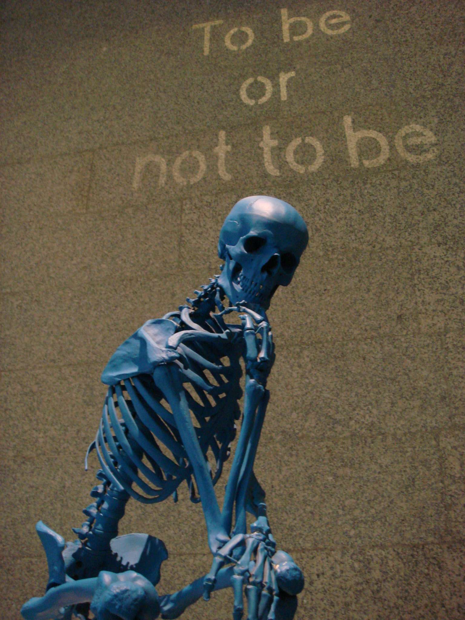 Blue skeletal sculpture posed in a contemplative seated position with one hand supporting its chin, evoking Rodin’s “The Thinker.” Faded text on the wall behind the sculpture reads “To be or not to be,” referencing Shakespeare’s Hamlet.
