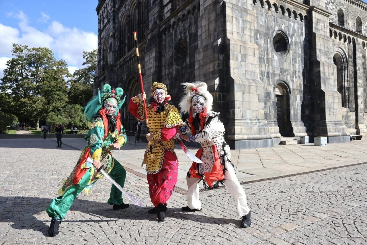 Three performers in colorful Chinese opera costumes and painted facial makeup pose dramatically with swords and a staff in front of the Lund Cathedral.