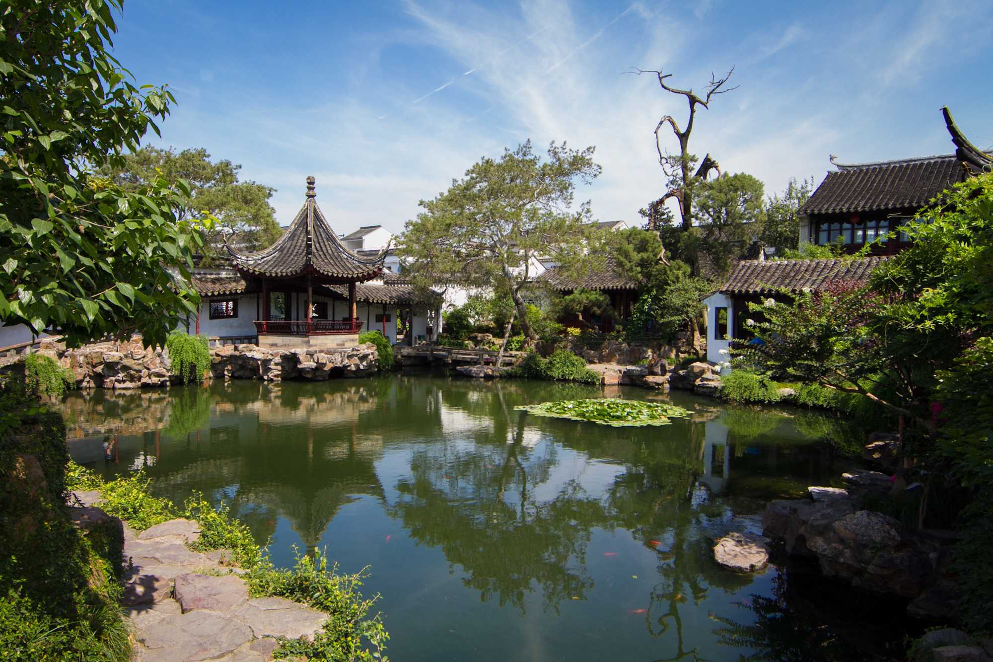 Traditional Chinese garden with a calm pond surrounded by trees, rocks, and classical pavilion architecture. Curved tiled rooftops and white-walled buildings reflect in the water beneath a bright blue sky with wispy clouds, while lily pads float near the center of the pond.