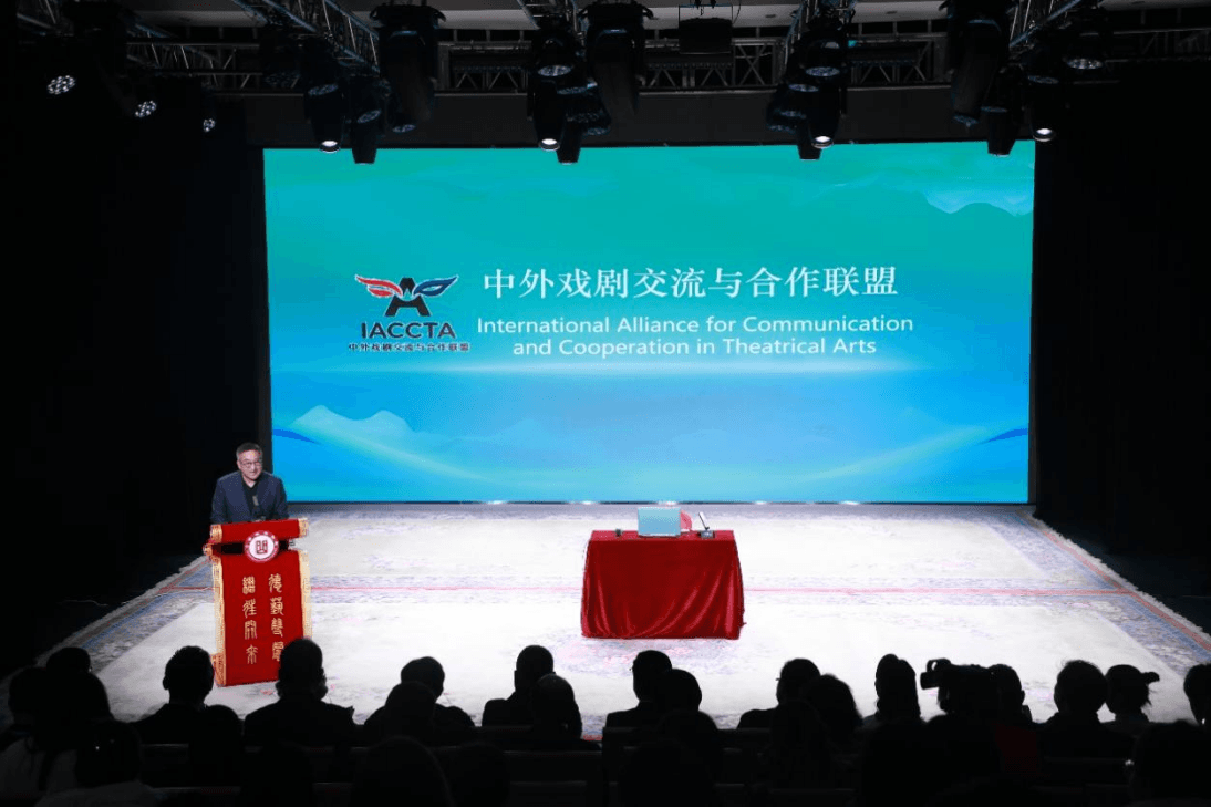 院长助理吴新苗在灯光明亮的舞台上主持中外戏剧交流与合作联盟(IACCTA)成立仪式。大屏幕呈青绿色背景,上面显示着该联盟的标志以及中英文名称。