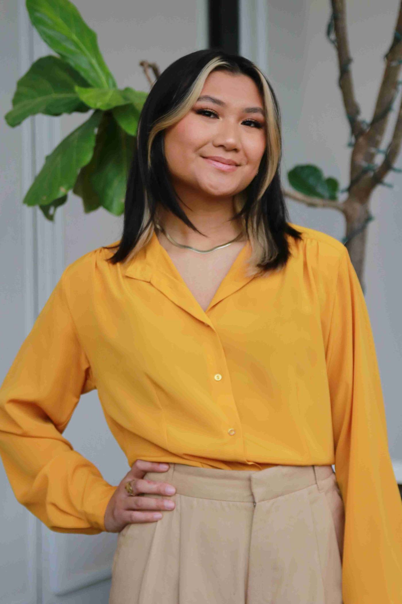 Portrait of person with long hair wearing a golden button up top smiling in front of a tree.