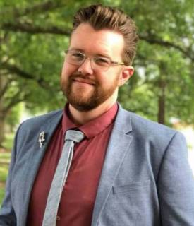 A picture of a white person smiling with facial hair and glasses dressed in formal attire with trees and greenery in the background.