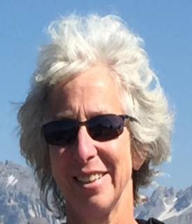 A picture of smiling white person with sunglasses and white hair with mountains and a blue sky in the background.