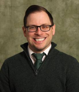 Portrait photo of a smiling white person with brown hair and glasses wearing a zip up fleece with a green tie underneath.