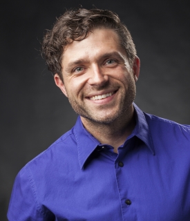A picture of a smiling white person with brown curly hair and a blue button up shirt with a black and gray background.