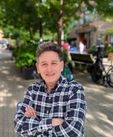 picture of a man with a flannel shirt on with a background blurred but can see some trees and walkway