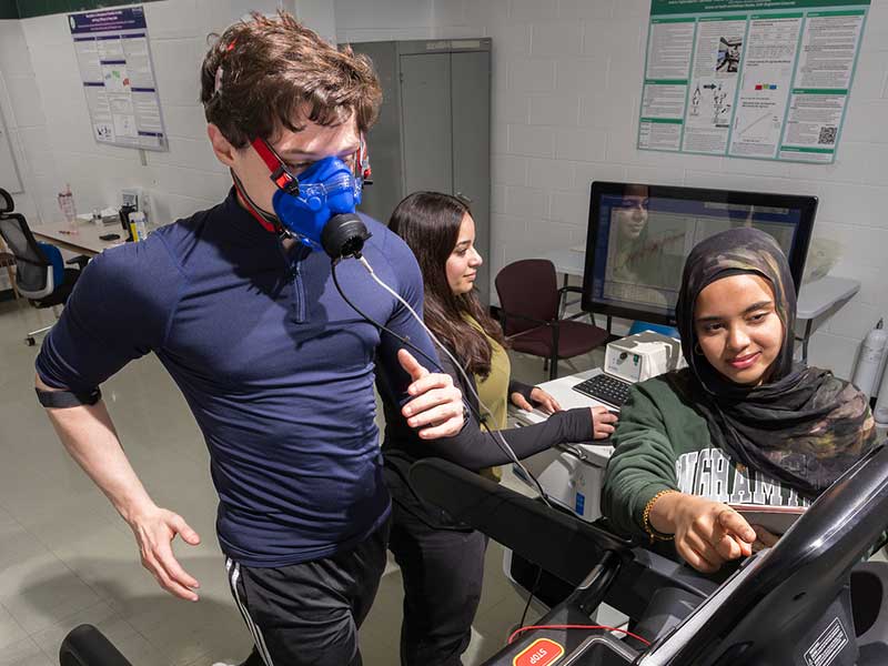 Student running on treadmill while one student works computer at nearby station and another controls treadmill