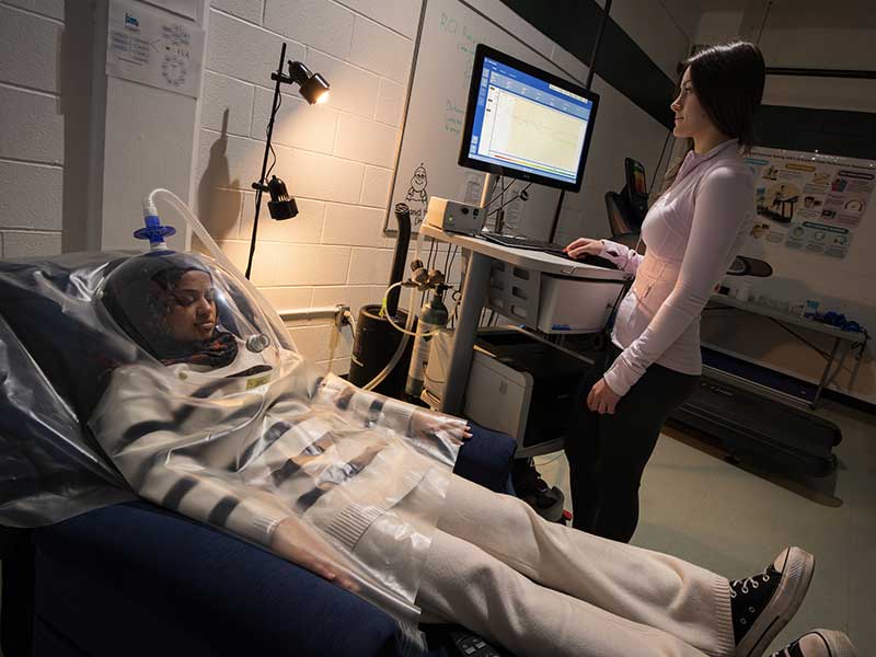 Student reclining in chair with testing hood while another student works controls at a nearby computer station