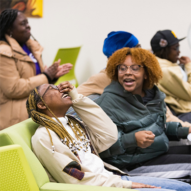 Two female students share a laugh while sitting on a couch.