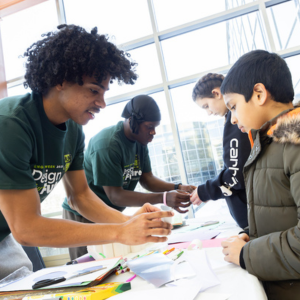 A Black male student demonstrates a task to a visiting youth.