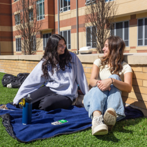 Two female students sit on the grass outside.