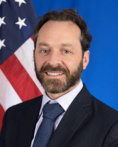 A man with a dark brown beard standing in front of an American flag