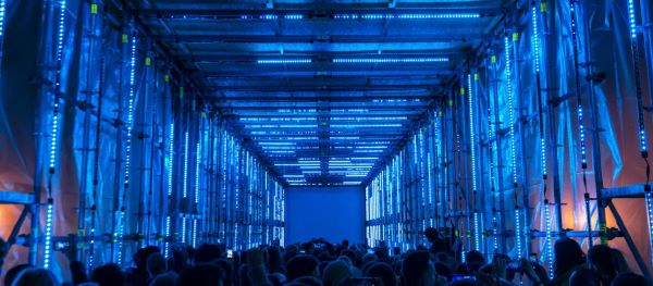 Crowd stands inside colorfully lighted scaffolding and watches projection.